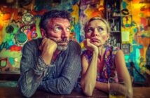 A man and woman sit side by side at a bar, both resting their heads on their hands and gazing in different directions with bored or thoughtful expressions. The background is colorful and abstract.