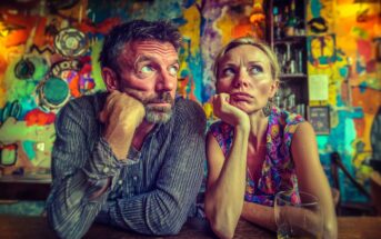 A man and woman sit side by side at a bar, both resting their heads on their hands and gazing in different directions with bored or thoughtful expressions. The background is colorful and abstract.