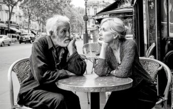 Two older adults sit at a small café table outside, resting their heads on their hands and gazing at each other, surrounded by a city street scene.