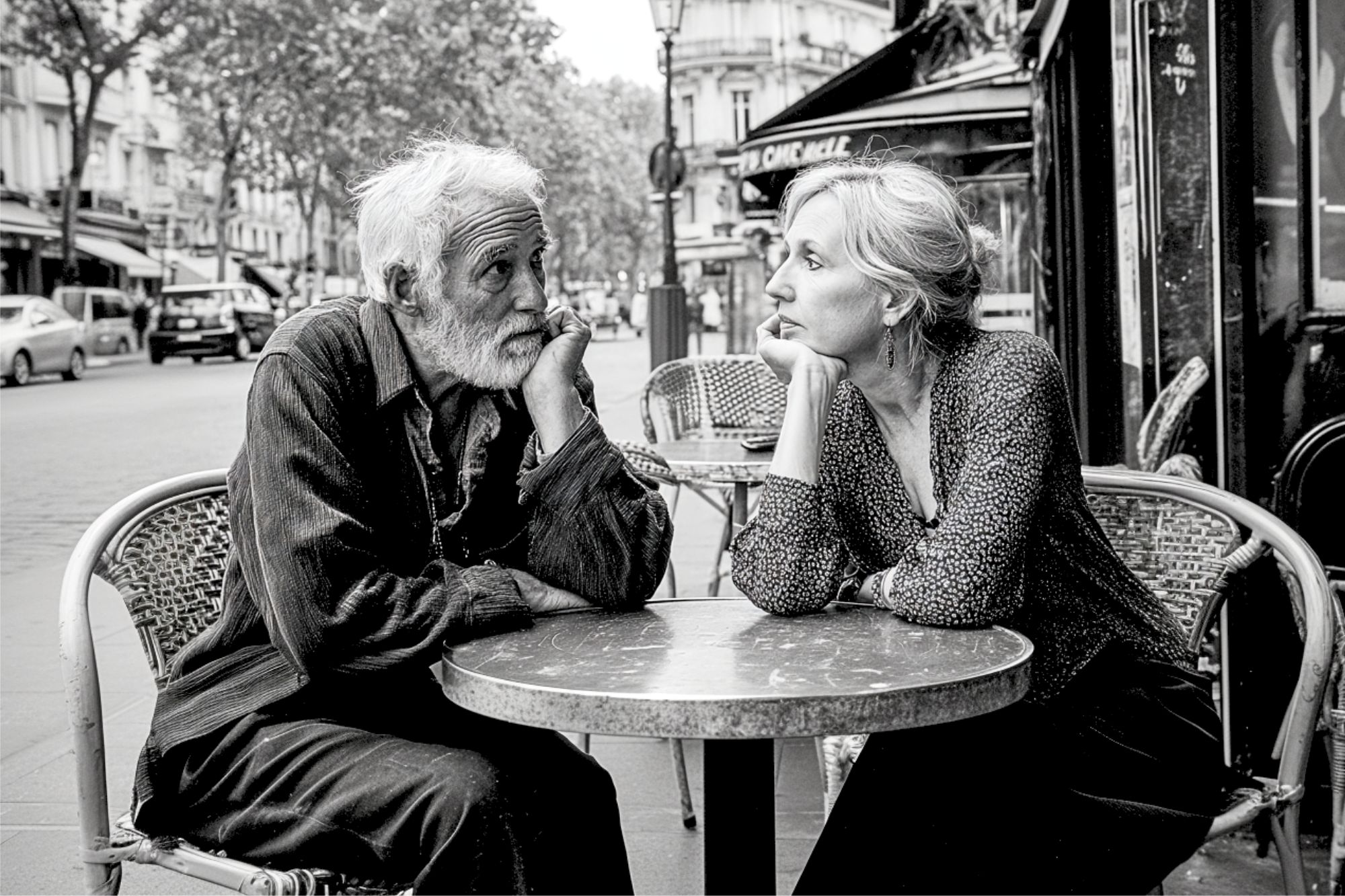 Two older adults sit at a small café table outside, resting their heads on their hands and gazing at each other, surrounded by a city street scene.