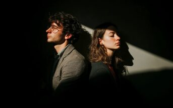 A man and a woman sit back-to-back in partial darkness, both facing away and looking serious. A beam of light highlights their faces, creating dramatic shadows on the wall behind them.