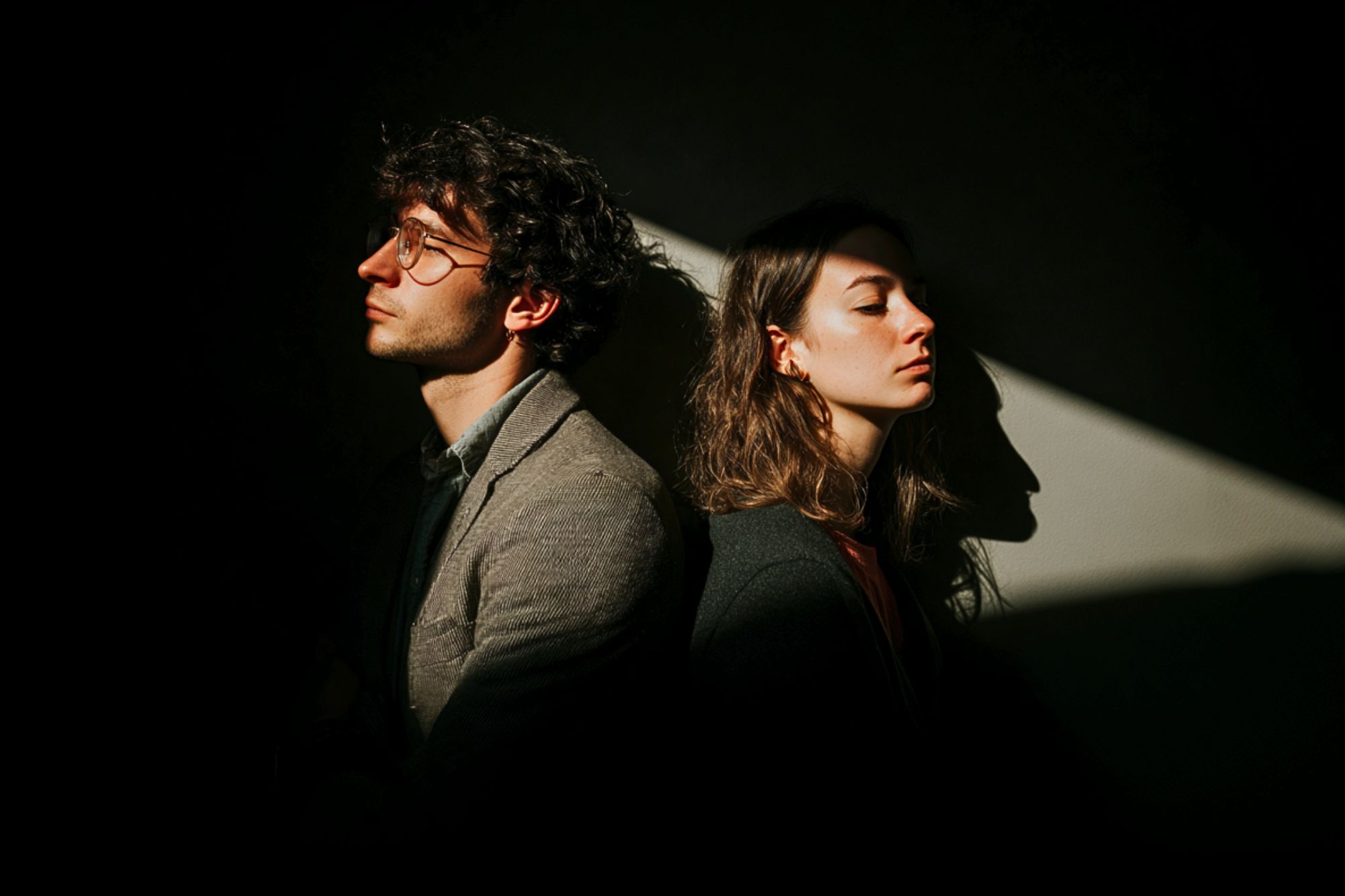 A man and a woman sit back-to-back in partial darkness, both facing away and looking serious. A beam of light highlights their faces, creating dramatic shadows on the wall behind them.