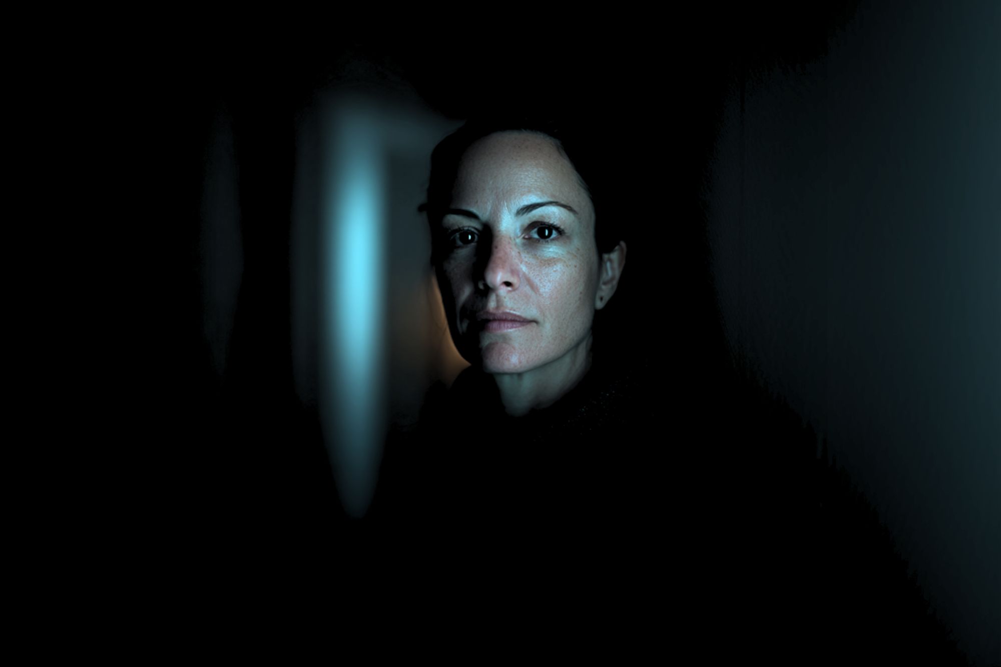A woman with a serious expression stands in a dimly lit hallway, her face illuminated by soft blue light, while the background fades into darkness.
