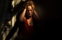A woman sits in a dimly lit room, holding her head in her hands with a distressed expression, partially illuminated by sunlight coming through a window.