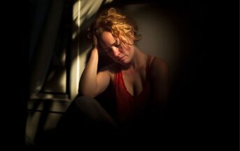 A woman with curly hair sits in a dark room, lit by sunlight through a window. She is wearing a red top, resting her head on her hand, with a thoughtful or tired expression. Shadows fall across her face and body.