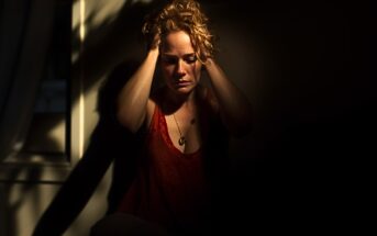A woman sits in a dimly lit room, holding her head in her hands with a distressed expression, partially illuminated by sunlight coming through a window.