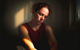 A woman with curly hair in a red sleeveless top sits in dim light, looking down with a somber expression. Sunlight creates a soft, warm highlight on her face and arm, with deep shadows around her.