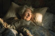 An older woman with gray hair lies in bed, smiling gently as warm sunlight shines on her face. She is surrounded by soft, floral-patterned bedding and appears peaceful and content.