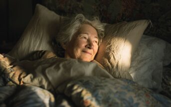 An older woman with gray hair lies in bed, smiling gently as warm sunlight shines on her face. She is surrounded by soft, floral-patterned bedding and appears peaceful and content.