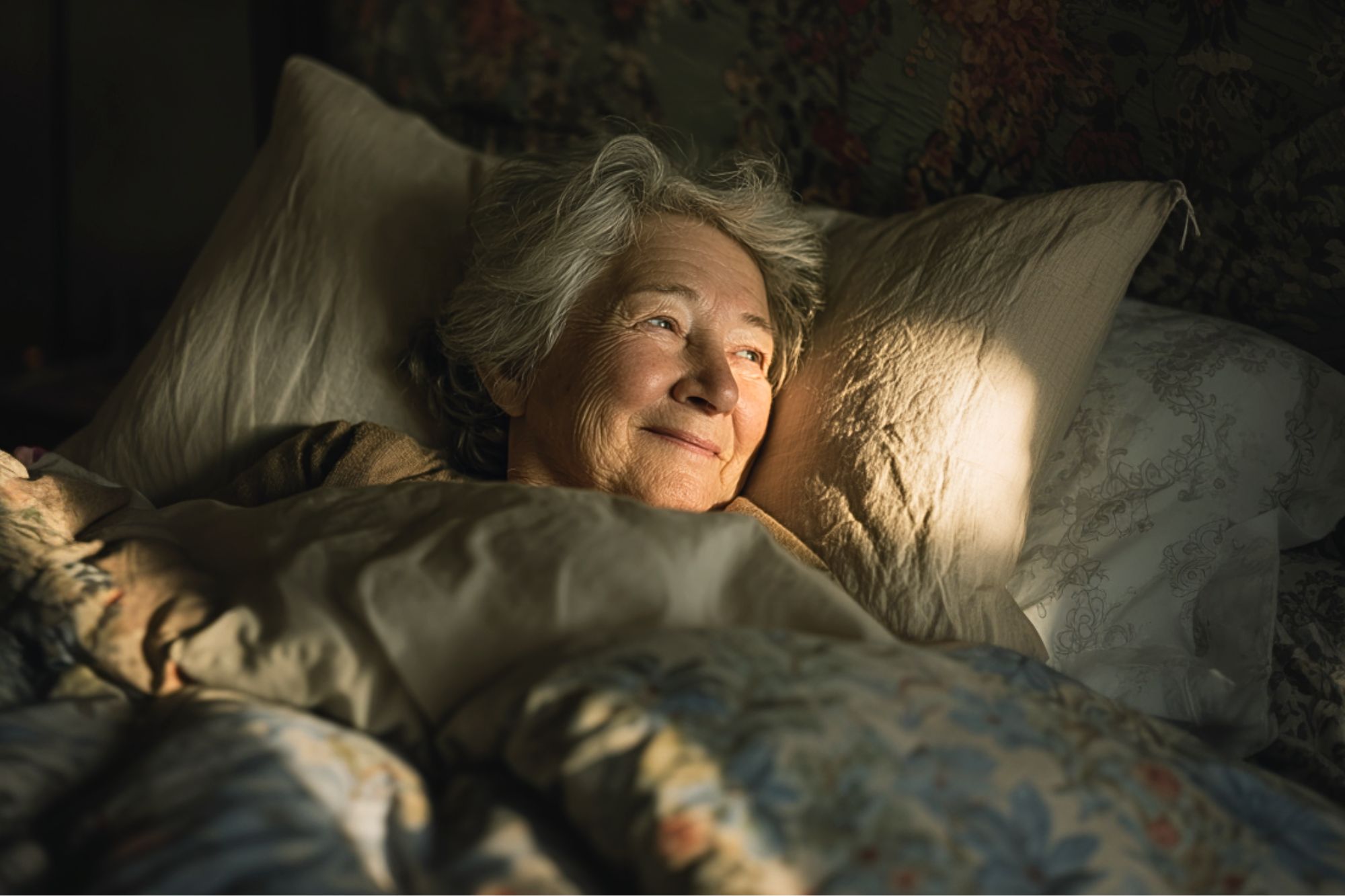 An older woman with gray hair lies in bed, smiling gently as warm sunlight shines on her face. She is surrounded by soft, floral-patterned bedding and appears peaceful and content.