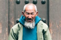 An elderly man with a long white beard and short gray hair wears a teal hoodie and a light green jacket, standing in front of a textured brown door, looking intently at the camera with a serious expression.
