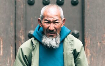 An elderly man with a long white beard and short gray hair wears a teal hoodie and a light green jacket, standing in front of a textured brown door, looking intently at the camera with a serious expression.