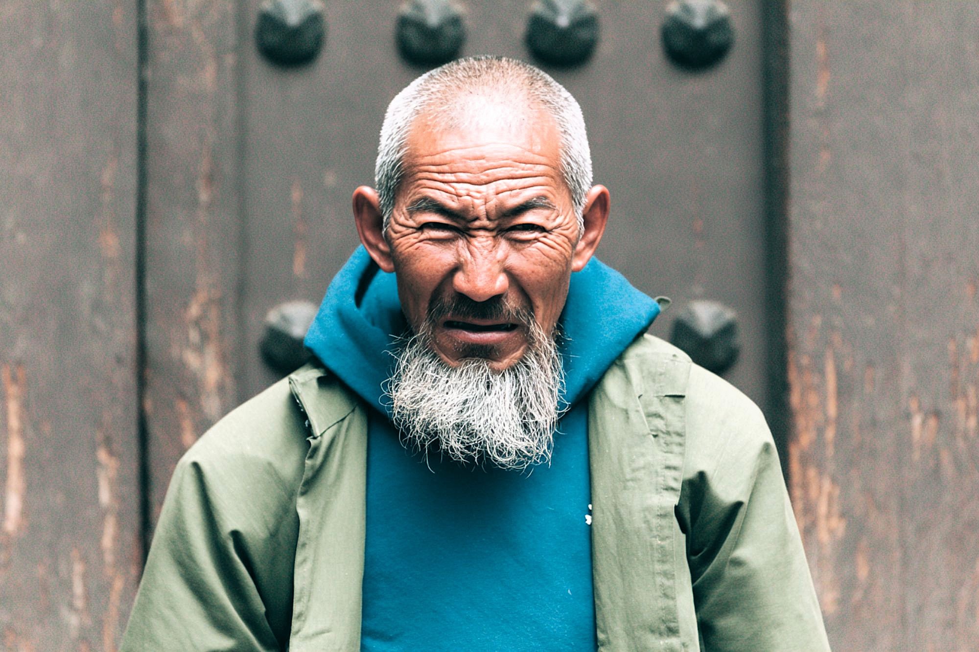 An elderly man with a long white beard and short gray hair wears a teal hoodie and a light green jacket, standing in front of a textured brown door, looking intently at the camera with a serious expression.