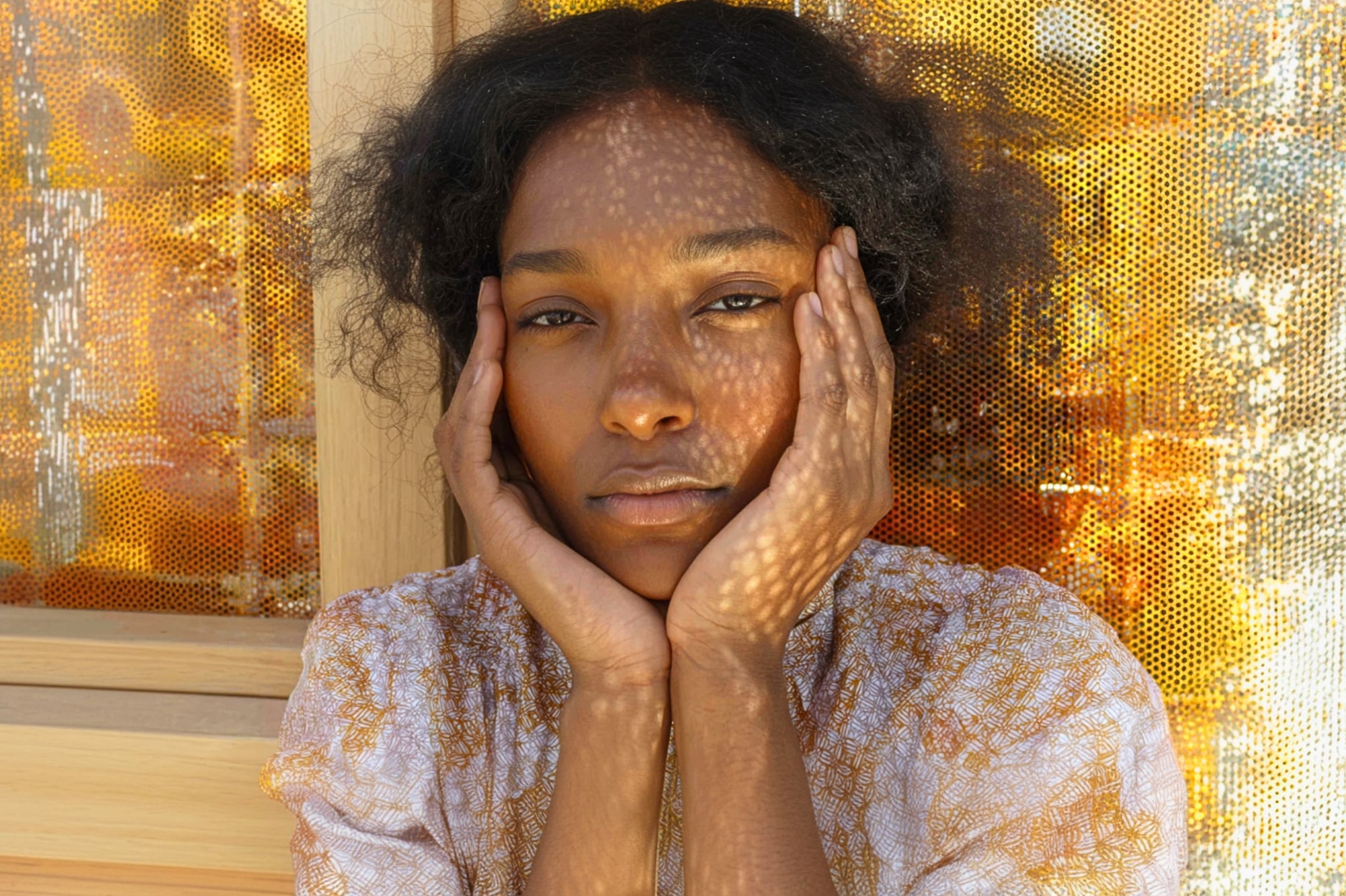 A person with medium-dark skin and curly hair rests their face in their hands, looking thoughtfully at the camera. Dappled sunlight creates patterned shadows on their face and background.