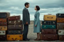 A man and woman in vintage coats stand facing each other on a rocky ground, separated by two tall stacks of old suitcases under a cloudy sky.