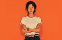 A woman with curly hair stands against an orange background, wearing a white t-shirt, black jeans, and an orange belt. She has her arms crossed and a neutral expression on her face.