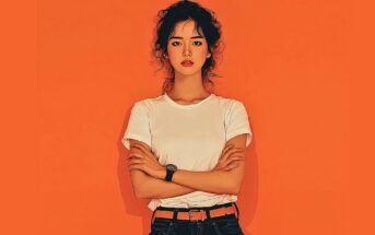 A woman with curly hair stands against an orange background, wearing a white t-shirt, black jeans, and an orange belt. She has her arms crossed and a neutral expression on her face.