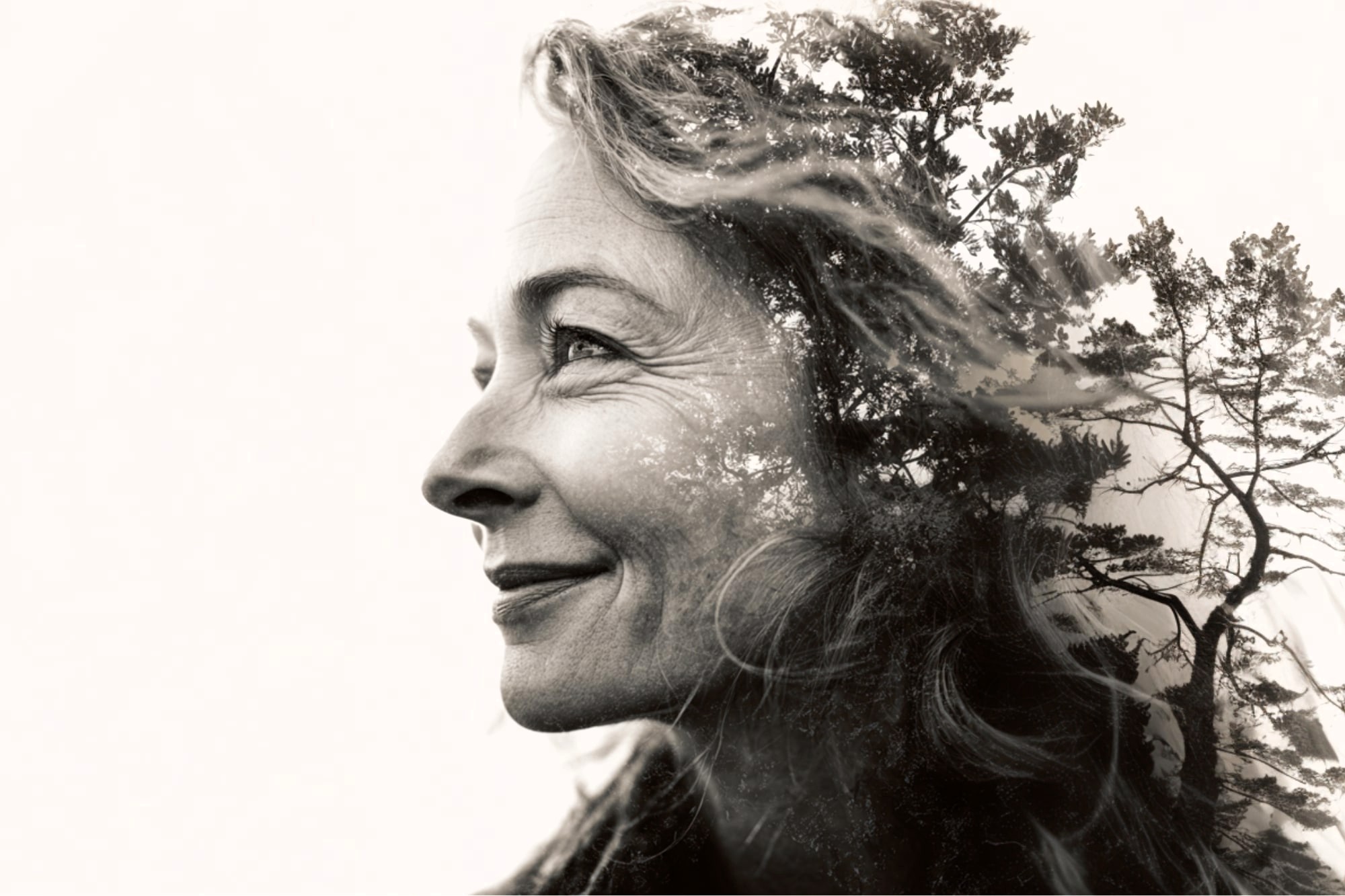 A black and white double exposure image of a smiling woman’s side profile blended with silhouettes of tall trees, creating an artistic and serene effect.
