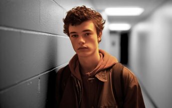 A teenage boy with curly hair, wearing a brown jacket and backpack, stands against a blue hallway wall under fluorescent lighting, looking thoughtfully at the camera.