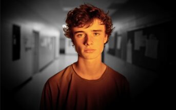 A teenage boy with curly hair stands in a dimly lit school hallway, looking seriously at the camera. The background is blurred, drawing focus to his expression.