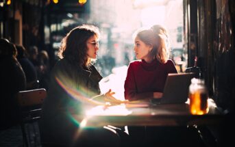 Two women sit at an outdoor café table, talking. One gestures while the other listens. Sunlight streams in, creating lens flare. A laptop and drink jar are on the table. The scene feels warm and casual.