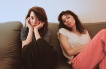 Two women sit on a couch indoors. One woman, with dark hair and striped shirt, rests her face in her hands and looks at the camera. The other, in a white t-shirt and pink pants, leans back with a neutral expression.