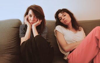 Two women sit on a couch indoors. One woman, with dark hair and striped shirt, rests her face in her hands and looks at the camera. The other, in a white t-shirt and pink pants, leans back with a neutral expression.