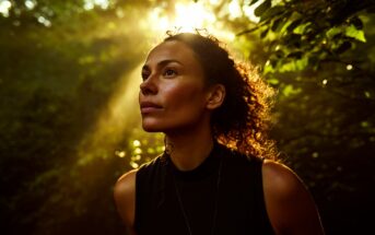 A woman with curly hair stands in a forest, looking thoughtfully upward as sunlight filters through the trees, illuminating her face and creating a warm, serene atmosphere.