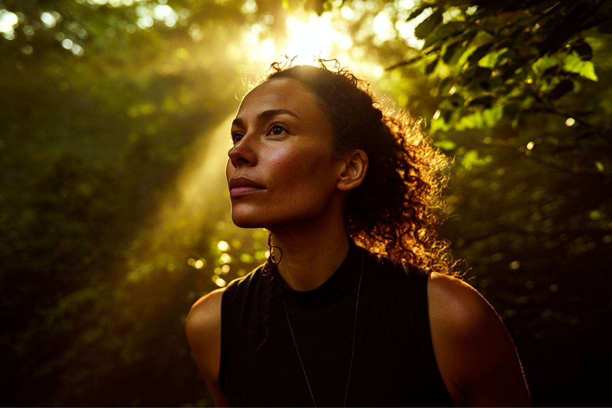 A woman with curly hair stands in a forest, looking thoughtfully upward as sunlight filters through the trees, illuminating her face and creating a warm, serene atmosphere.