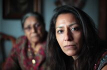 A woman with long dark hair looks seriously at the camera, while an older woman sits blurred in the background. Both wear patterned clothing in a dimly lit room.