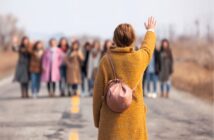 A person in a yellow coat and pink backpack stands on a road, waving to a group of people in the distance who appear to be waving back. The scene is outdoors on a cloudy day.