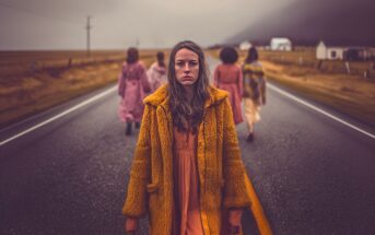 A young woman in an orange coat stands on a rural road, looking at the camera, while four other women in dresses walk away from her toward distant houses under a cloudy, moody sky.
