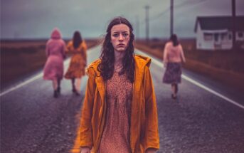 A young woman in an orange coat stands on a wet road, looking forward. Three women in dresses walk away from her, and a small house is visible on the roadside under a cloudy sky.