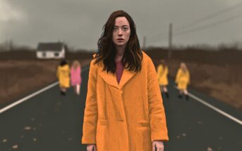 A woman in an orange coat stands in the middle of a road, with four other people walking away from her into the distance.