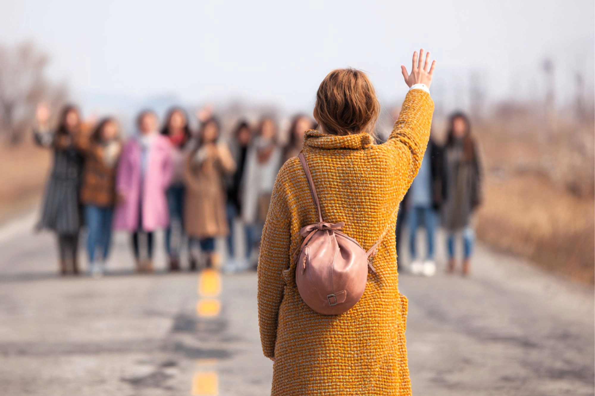 A person in a yellow coat and pink backpack stands on a road, waving to a group of people in the distance who appear to be waving back. The scene is outdoors on a cloudy day.