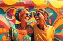 Two older women with gray hair, colorful headbands, and sunglasses laugh together in front of a vibrant, abstract mural with vivid orange, yellow, blue, and pink patterns.