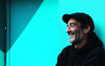 A middle-aged man with a beard wearing a black hoodie and cap smiles while leaning against a turquoise geometric-patterned wall.