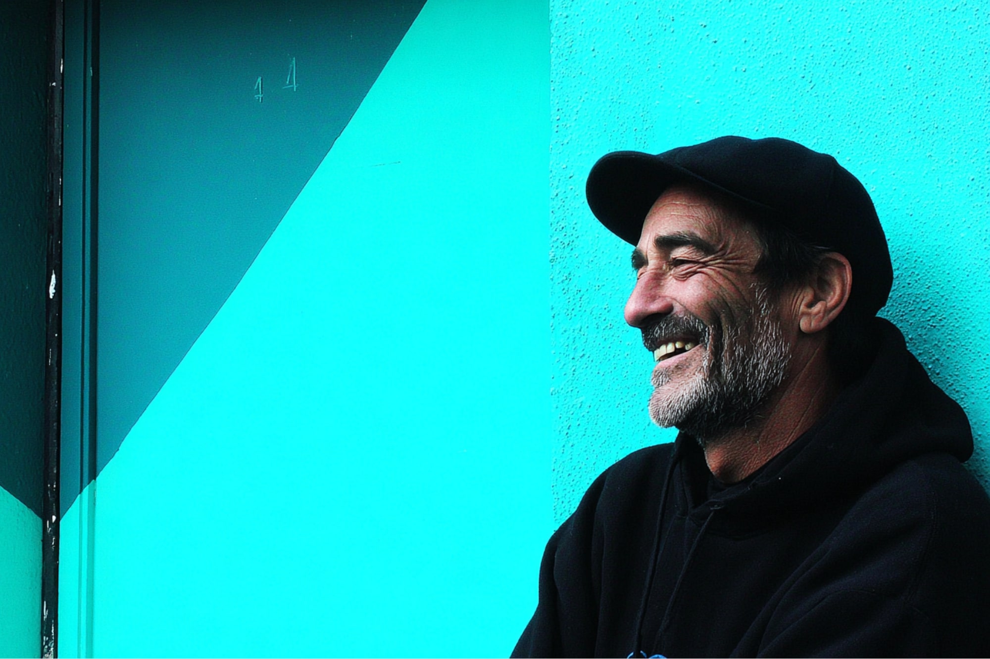 A middle-aged man with a beard wearing a black hoodie and cap smiles while leaning against a turquoise geometric-patterned wall.
