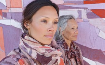 Two women, one younger and one older, stand in front of a colorful abstract mural. Both wear patterned scarves and look thoughtfully into the distance. The mural features shades of pink, purple, orange, and white.