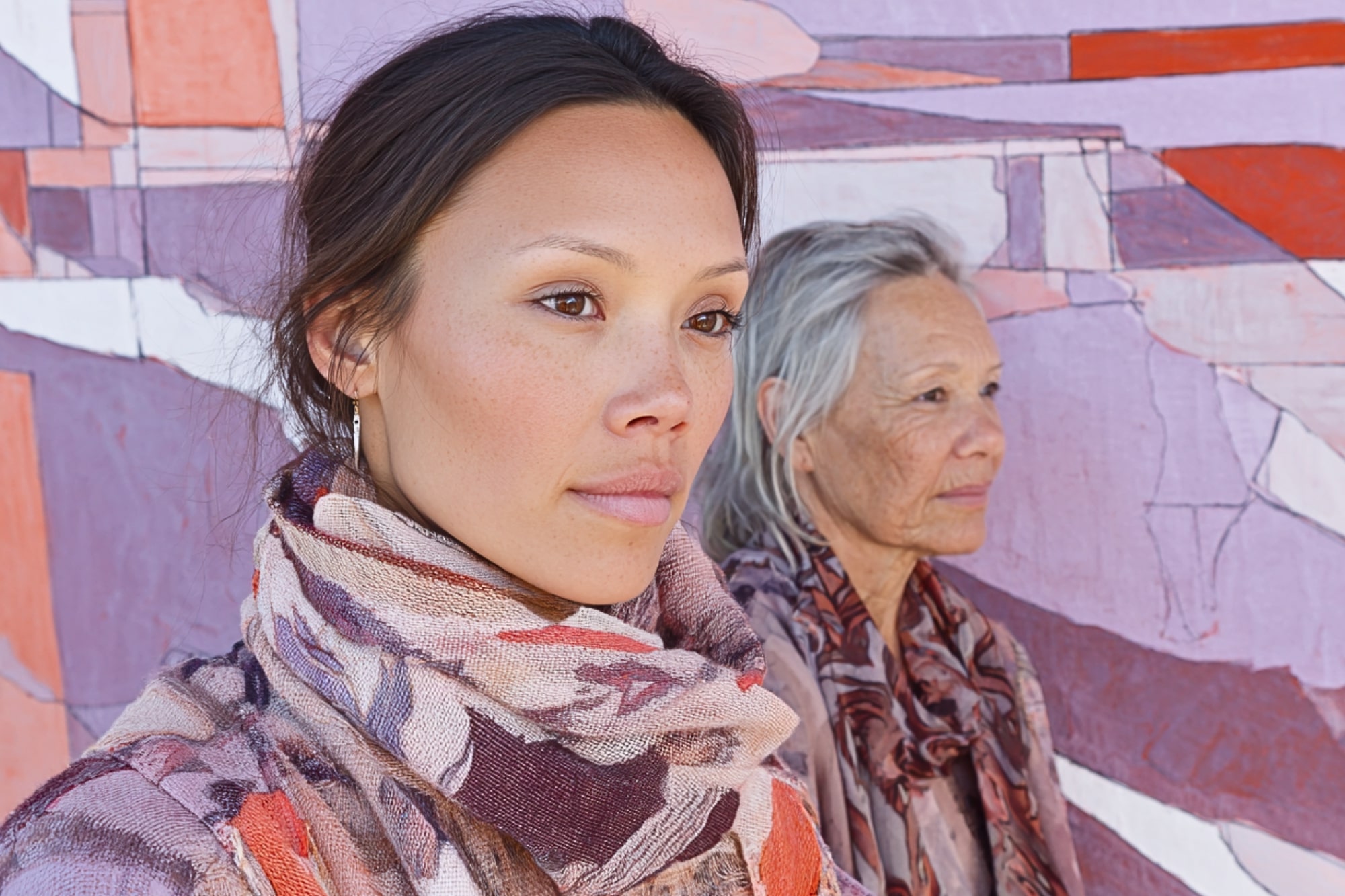 Two women, one younger and one older, stand in front of a colorful abstract mural. Both wear patterned scarves and look thoughtfully into the distance. The mural features shades of pink, purple, orange, and white.