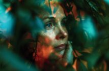 A woman’s thoughtful face is partially obscured by out-of-focus green leaves, with warm orange and cool blue lighting creating a dreamy, contemplative atmosphere. She rests her chin on her hand and gazes upward.
