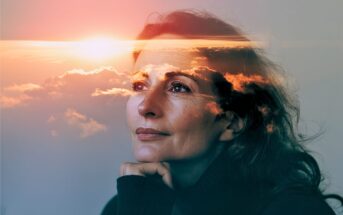 A woman gazes thoughtfully upward, her chin resting on her hand. A double exposure effect overlays her face with a vibrant sunset and clouds, creating a dreamy and reflective atmosphere.