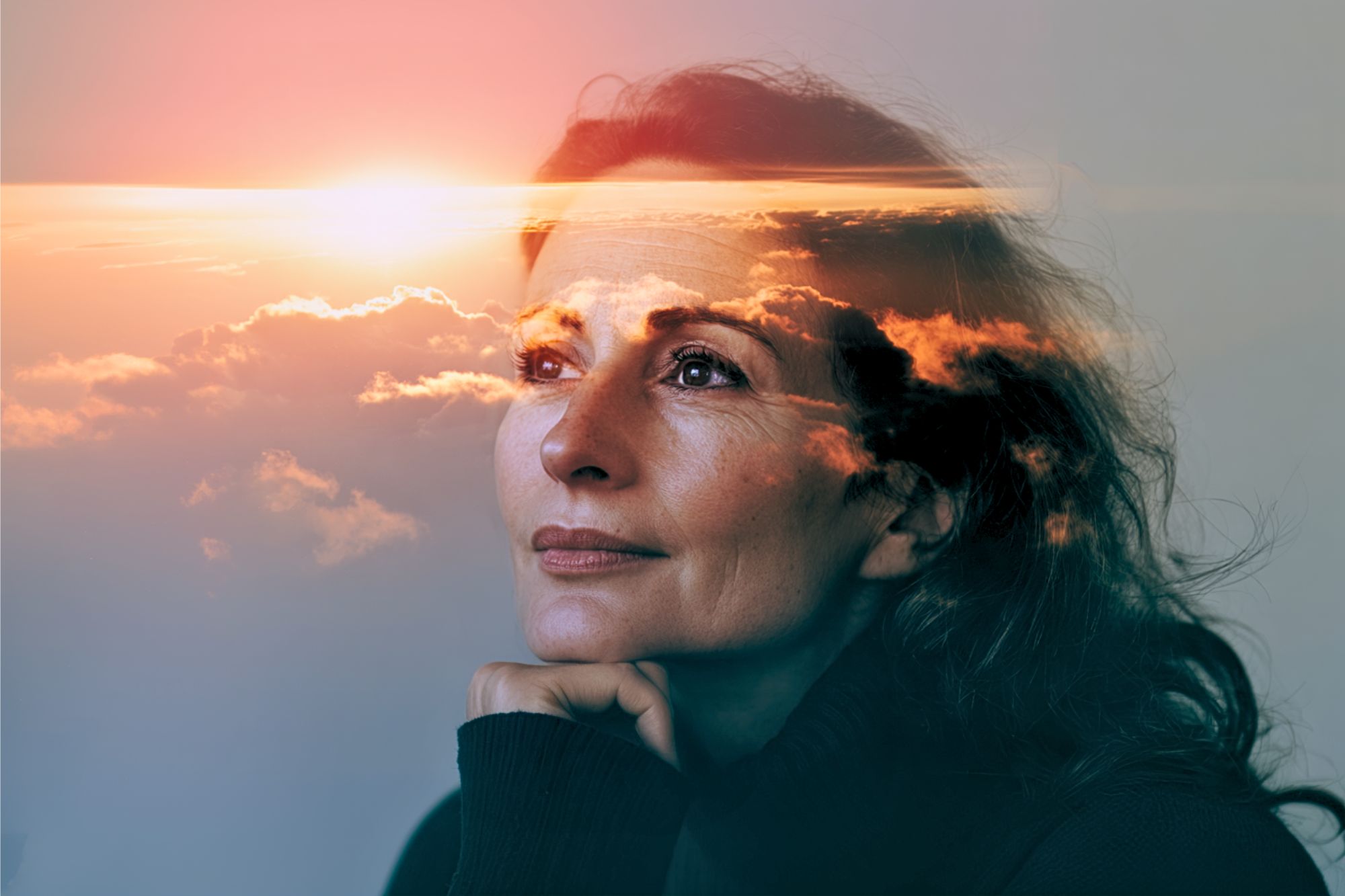 A woman gazes thoughtfully upward, her chin resting on her hand. A double exposure effect overlays her face with a vibrant sunset and clouds, creating a dreamy and reflective atmosphere.