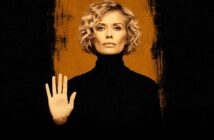 A woman with short, curly blonde hair and a serious expression faces forward, wearing a black turtleneck and holding up her left hand in a stop gesture. The background is black with a vertical orange stripe behind her.