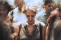 A woman with short hair stands looking down, appearing pensive, surrounded by a blurred crowd in motion, creating a sense of isolation and introspection amid busy surroundings.
