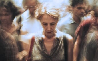 A woman with short hair stands looking down, appearing pensive, surrounded by a blurred crowd in motion, creating a sense of isolation and introspection amid busy surroundings.