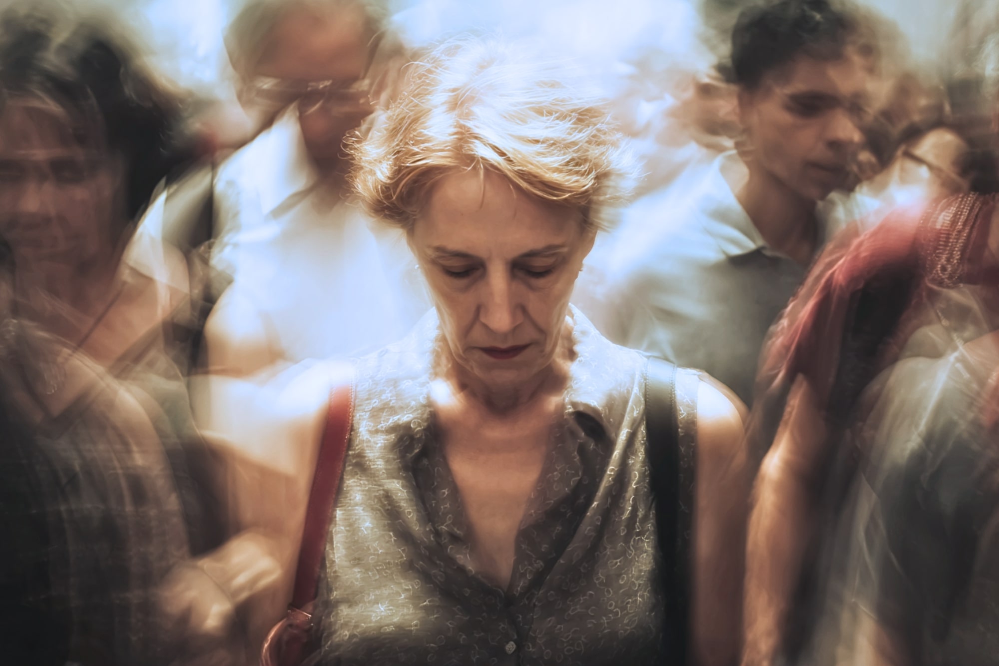 A woman with short hair stands looking down, appearing pensive, surrounded by a blurred crowd in motion, creating a sense of isolation and introspection amid busy surroundings.