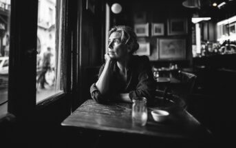 A woman sits alone at a table in a dimly lit café, gazing thoughtfully out the window. The scene is in black and white, evoking a contemplative or nostalgic mood.