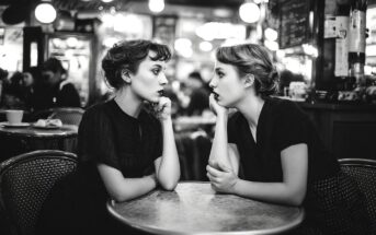 Two young women sit across from each other at a round café table, gazing intently at each other in a busy, softly lit café. The image is in black and white, creating a nostalgic, vintage atmosphere.