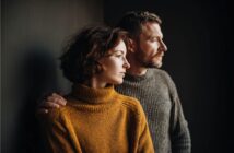 A woman in a mustard-colored sweater and a man in a gray sweater stand close together, looking thoughtfully out of frame toward the light. The man's arm rests gently on the woman's shoulder.